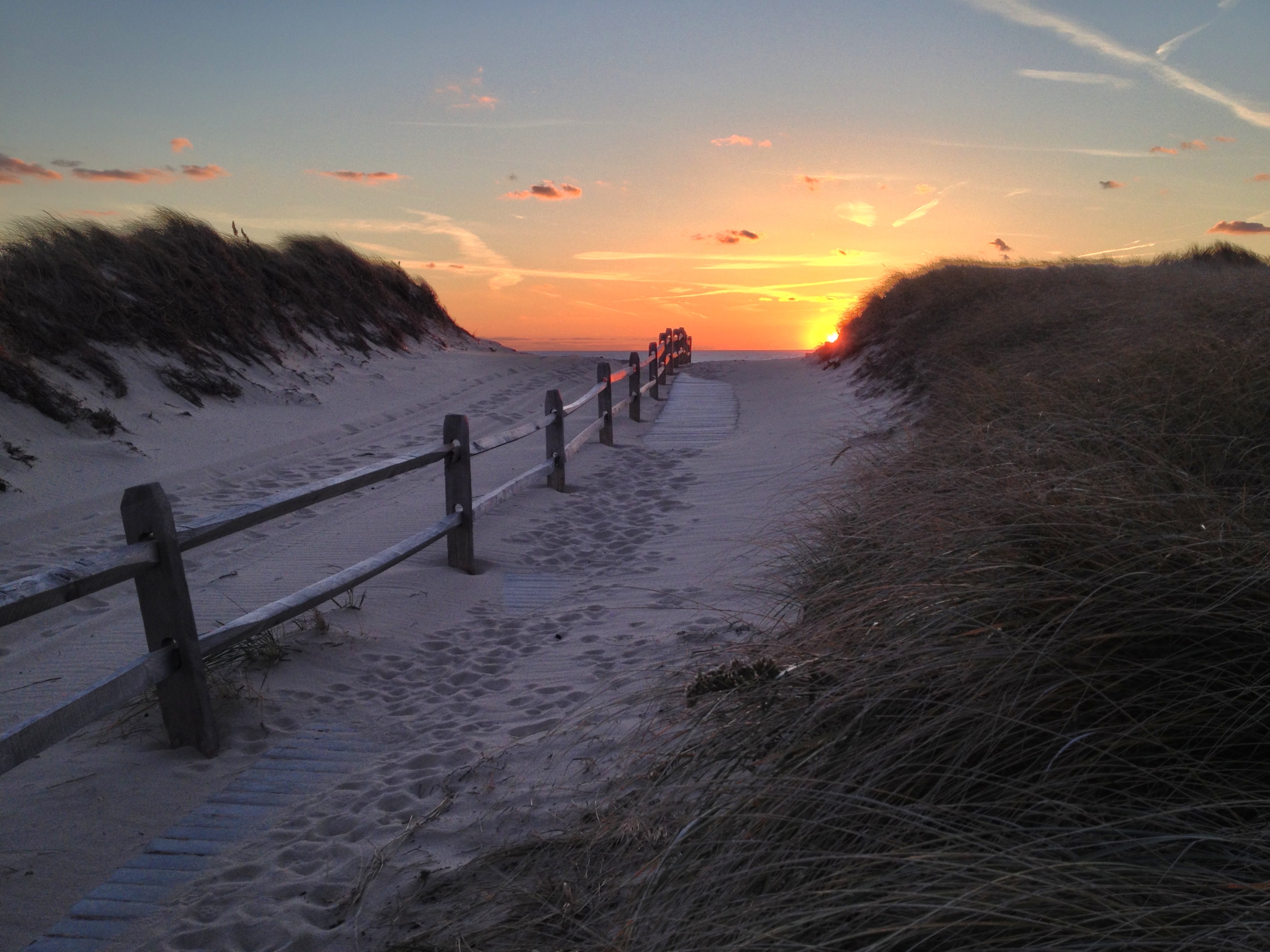 sand covered path heading toward the beach at sunset with beach grass on either side of the path and a fence in the center